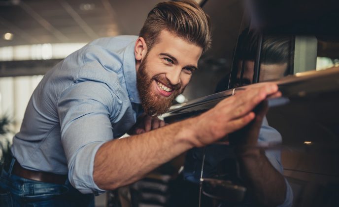 Ein junger Mann begutachtet freudig die sauberen Reflexionen in der Karosserie seines Autos und streicht dabei mit seiner Hand über den schwarzen Lack.